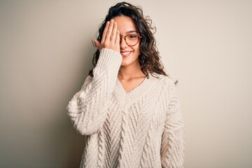 Beautiful woman with curly hair wearing casual sweater and glasses over white background covering one eye with hand, confident smile on face and surprise emotion.