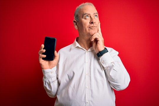 Middle age hoary man holding smartphone showing screen over isolated red background serious face thinking about question, very confused idea