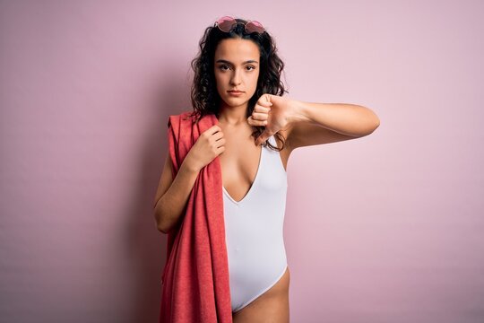 Beautiful woman with curly hair on vacation wearing white swimsuit holding beach towel with angry face, negative sign showing dislike with thumbs down, rejection concept