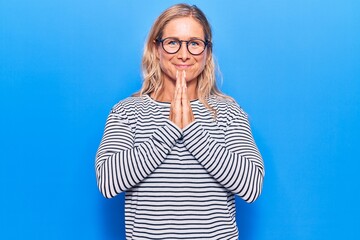 Obraz na płótnie Canvas Middle age caucasian blonde woman wearing casual striped sweater and glasses praying with hands together asking for forgiveness smiling confident.