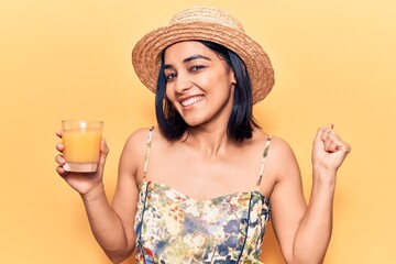 Young beautiful latin woman wearing summer hat drinking glass of orange juice screaming proud, celebrating victory and success very excited with raised arm