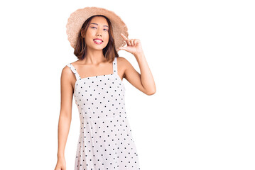 Young beautiful chinese girl wearing summer hat smiling and confident gesturing with hand doing small size sign with fingers looking and the camera. measure concept.