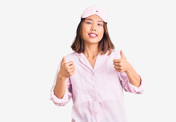 Young beautiful chinese girl wearing sleep mask and pajama success sign doing positive gesture with hand, thumbs up smiling and happy. cheerful expression and winner gesture.