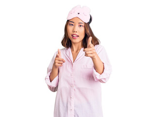 Young beautiful chinese girl wearing sleep mask and pajama pointing fingers to camera with happy and funny face. good energy and vibes.