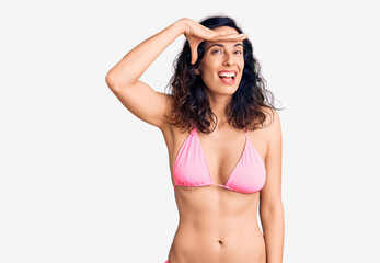 Young beautiful hispanic woman wearing bikini very happy and smiling looking far away with hand over head. searching concept.