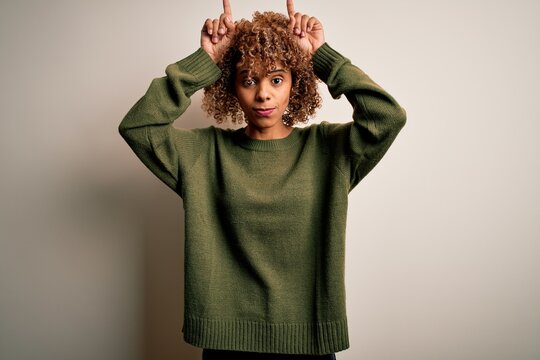 Beautiful african american woman with curly hair wearing casual sweater over white background doing funny gesture with finger over head as bull horns