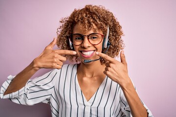 African american curly call center agent woman working using headset over pink background smiling cheerful showing and pointing with fingers teeth and mouth. Dental health concept.