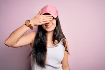 Young brunette woman wearing casual sport cap over pink background smiling and laughing with hand on face covering eyes for surprise. Blind concept.