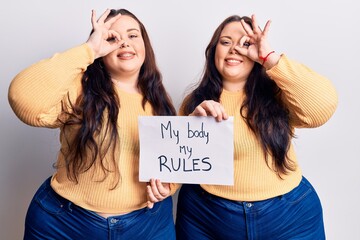 Young plus size twins holding my body my rules banner smiling happy doing ok sign with hand on eye looking through fingers