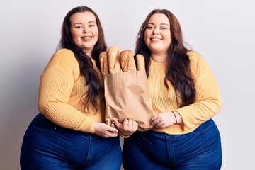 Young plus size twins holding paper bag with bread looking positive and happy standing and smiling with a confident smile showing teeth