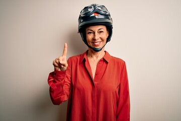 Middle age motorcyclist woman wearing motorcycle helmet over isolated white background showing and pointing up with finger number one while smiling confident and happy.