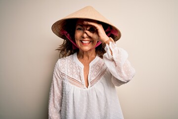Middle age brunette woman wearing asian traditional conical hat over white background doing ok gesture with hand smiling, eye looking through fingers with happy face.