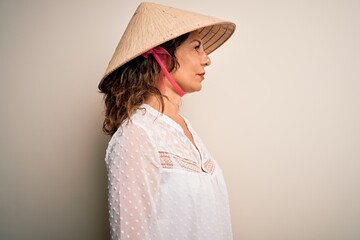 Middle age brunette woman wearing asian traditional conical hat over white background looking to side, relax profile pose with natural face with confident smile.