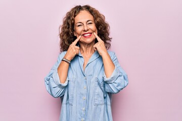 Middle age beautiful woman wearing casual denim shirt standing over pink background Smiling with open mouth, fingers pointing and forcing cheerful smile