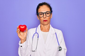 Middle age senior cardiologist doctor woman holding red heart over purple background with a...