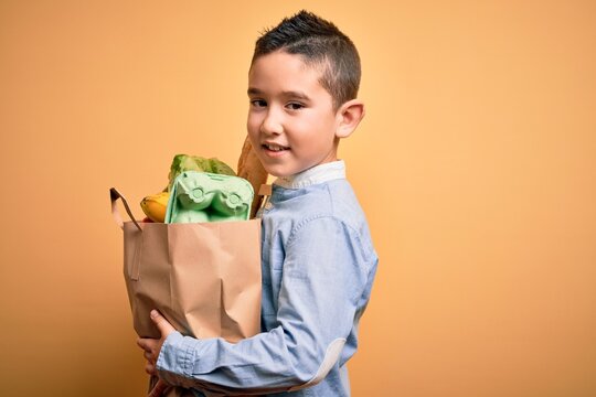 Adorable toddler holding paper bag with food standing over isolated yellow background
