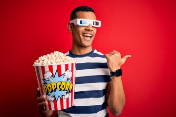 Young handsome african american man watching movie using 3d glasses eating popcorns smiling with happy face looking and pointing to the side with thumb up.