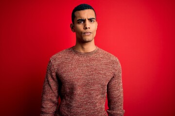 Young handsome african american man wearing casual sweater standing over red background skeptic and...