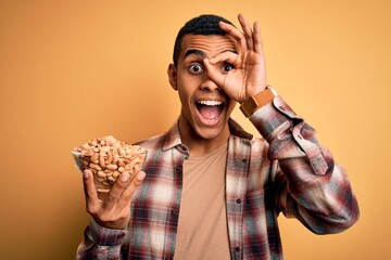 Handsome african american man holding bowl with heathy peanuts over yellow background with happy face smiling doing ok sign with hand on eye looking through fingers