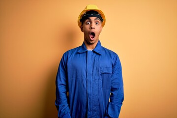 Young handsome african american worker man wearing blue uniform and security helmet afraid and shocked with surprise and amazed expression, fear and excited face.