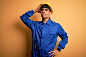 Young handsome african american worker man wearing blue uniform and security helmet confuse and wondering about question. Uncertain with doubt, thinking with hand on head. Pensive concept.
