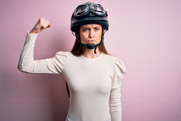 Young beautiful motorcyclist woman with blue eyes wearing moto helmet over pink background Strong person showing arm muscle, confident and proud of power