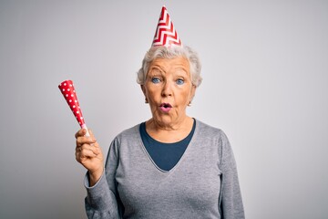 Senior beautiful grey-haired woman celebrating birthday wearing funny hat holding trumpet scared in shock with a surprise face, afraid and excited with fear expression