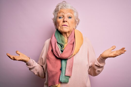 Senior beautiful grey-haired woman wearing casual sweater and scarf over pink background clueless and confused expression with arms and hands raised. Doubt concept.