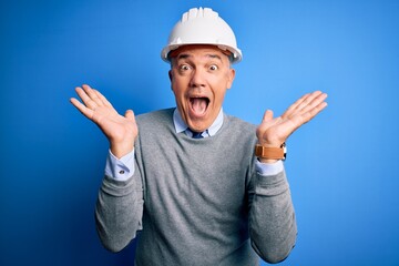Middle age handsome grey-haired engineer man wearing safety helmet over blue background celebrating crazy and amazed for success with arms raised and open eyes screaming excited. Winner concept
