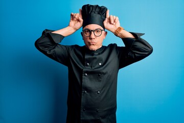 Middle age handsome grey-haired chef man wearing cooker uniform and hat doing funny gesture with finger over head as bull horns