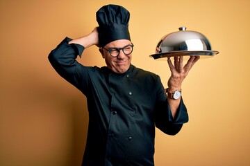 Middle age handsome grey-haired waiter man wearing cooker uniform and hat holding tray confuse and wondering about question. Uncertain with doubt, thinking with hand on head. Pensive concept.