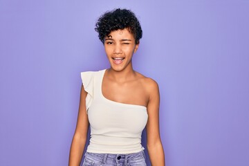 Young beautiful african american afro woman wearing casual t-shirt over purple background winking looking at the camera with sexy expression, cheerful and happy face.