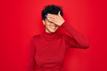Young beautiful african american afro woman wearing turtleneck sweater and glasses smiling and laughing with hand on face covering eyes for surprise. Blind concept.