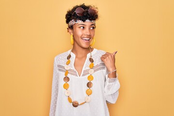 Young beautiful african american afro hippie woman wearing sunglasses and accessories smiling with happy face looking and pointing to the side with thumb up.