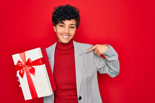 Beautiful african american afro business woman holding gift over isolated red background with surprise face pointing finger to himself