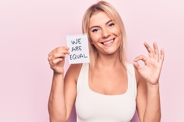 Young beautiful blonde woman asking for equality holding paper with we are equal message doing ok sign with fingers, smiling friendly gesturing excellent symbol