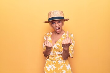 Beautiful blonde woman on vacation wearing summer hat and dress over yellow background Showing middle finger doing fuck you bad expression, provocation and rude attitude. Screaming excited