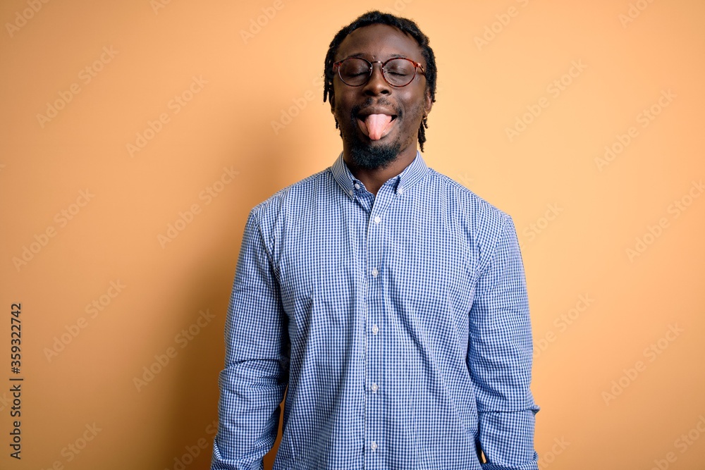 Sticker Young handsome african american man wearing shirt and glasses over yellow background sticking tongue out happy with funny expression. Emotion concept.