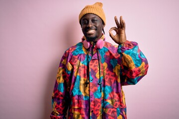 Young handsome african american man wearing colorful coat and cap over pink background smiling positive doing ok sign with hand and fingers. Successful expression.