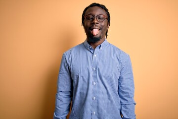 Fototapeta premium Young handsome african american man wearing shirt and glasses over yellow background sticking tongue out happy with funny expression. Emotion concept.