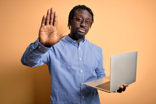 Young african american worker man working using laptop standing over yellow background with open hand doing stop sign with serious and confident expression, defense gesture