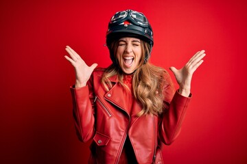 Young beautiful brunette motrocyclist woman wearing moto helmet over red background celebrating mad...