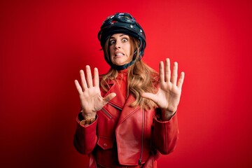 Young beautiful brunette motrocyclist woman wearing moto helmet over red background afraid and terrified with fear expression stop gesture with hands, shouting in shock. Panic concept.