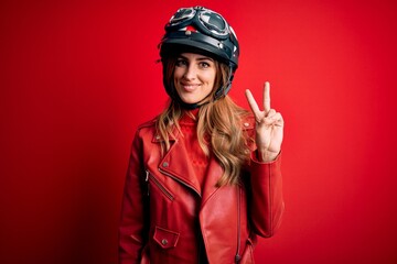 Young beautiful brunette motrocyclist woman wearing moto helmet over red background showing and pointing up with fingers number two while smiling confident and happy.