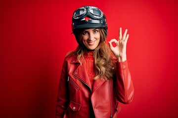 Young beautiful brunette motrocyclist woman wearing moto helmet over red background smiling positive doing ok sign with hand and fingers. Successful expression.