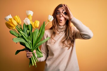 Young beautiful brunette woman holding bouquet of yellow tulips over isolated background doing ok gesture shocked with surprised face, eye looking through fingers. Unbelieving expression.