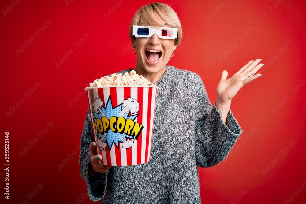 Wall mural Young blonde woman with short hair wearing 3d movie glasses and eating popcorn at the cinema very happy and excited, winner expression celebrating victory screaming with big smile and raised hands