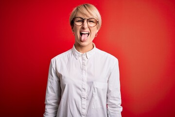 Young blonde business woman with short hair wearing glasses over red background sticking tongue out happy with funny expression. Emotion concept.