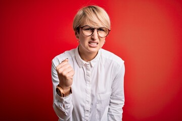 Young blonde business woman with short hair wearing glasses over red background angry and mad raising fist frustrated and furious while shouting with anger. Rage and aggressive concept.