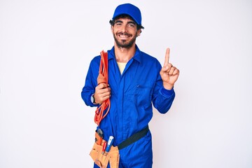 Handsome young man with curly hair and bear wearing builder jumpsuit uniform and electric cables smiling with an idea or question pointing finger with happy face, number one